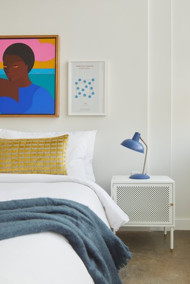 Modern bedroom with white nightstand, blue lamp, yellow pillow, eclectic art on wall.